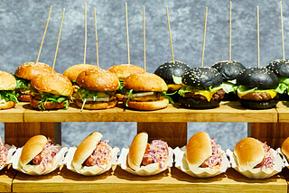 There are two wooden shelves of burgers and hotdogs. On the top shelf, on the left are chicken burgers consisting of a sesame seed bun, split in two, each containing a breaded, fried chicken fillet, a slice of cheese and lettuce. On the right of the top shelf are black charcoal buns, each containing a beef patty topped with a slice of melted cheese and some lettuce. The bottom shelf contains split hot dog buns, filled with hotdogs and pink coleslaw, each in a little paper tray.