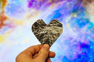 A hand holding up a heart-shaped piece of wrinkled gold foil against a pink, purple, blue, and gold background