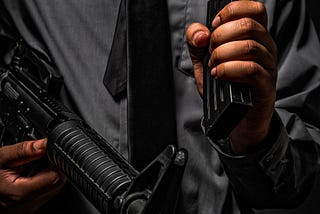 A man in a dark grey shirt and black tie holding an assault rifle and high capacity magazine