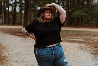 A fat, female-bodied human stands defiantly proud with one hand on their hip and the other hand atop their hat. They’re wearing a black top and tight jeans which show off their figure.