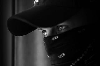 Man with piercing eyes wearing a Yankee hat and black bandana covering his face.