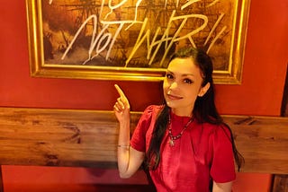 Woman in red shirt sitting at wooden table, smiling and pointing up at framed golden artwork behind her that reads ‘Make Cocktails Not War.’
