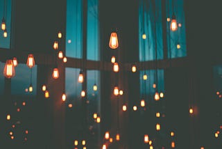 A picture of many hanging lightbulbs burning brightly against the backdrop of a dark room partially illuminated by light coming through blue curtains.