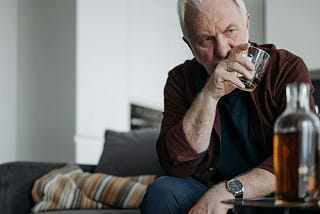Image shows older man looking frustrated and upset while drinking whiskey from a tumbler while bottle of whiskey sits on a table in the foreground.