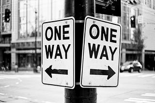 Two “One Way” traffic signs pointing in opposite directions on the corner of a city street.