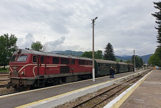 red train engine with green carriages sitting at quiet train platform under cloudy skies