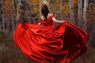 A woman in a red ball gown and gold crown, dancing in front of birch trees in the fall.