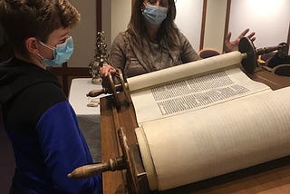 The young man reading from the Torah is Jack H. He is preparing for his Bar Mitzvah. The woman holding the Torah is Kim B., one of his teachers. The place is the chapel of Temple Shalom, where the Torah rests in the ark.