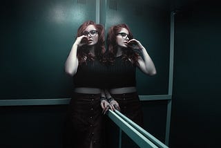 A woman leaning against the corner of an elevator. She is reflected in the mirrored side wall. She has red hair, and is wearing glasses, a black halter top short enough to reveal a little midriff, and a long, brown, buttoned skirt. She has one hand raised anxiously to her slightly parted lips.