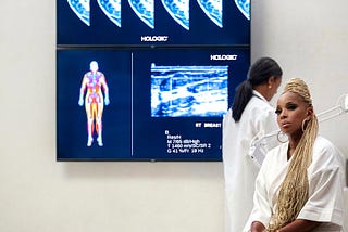 Photograph of a large doctor’s office with Mary J. Blige sitting in the foreground and body/breast imaging on the wall.