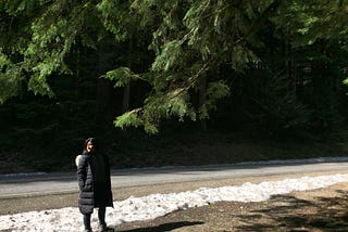 A woman stands on the side of the road with snow nearby, with the branches of an evergreen tree above her.
