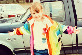 A photo from around 1995 of the author Krystal as a little girl wearing a rainbow windbreaker, a white shirt with rainbow hearts, jeans, and a backpack. She stands in front of a Ford Bronco smiling with her arms outstreched as if to say “come at me”.