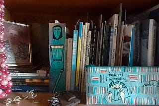 Bookshelf with photos, a bookmark, books, bits of jewelry, and a pencil case that says: fuck off I’m reading.