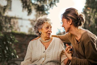 A woman talking with her elderly mother while smiling and putting an arm on the mother’s shoulder.