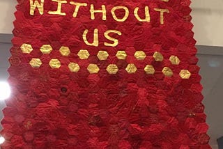 A banner made up of hundred of individually stitched red hexagons. In gold writting over the top it says Nothing About Us Without Us