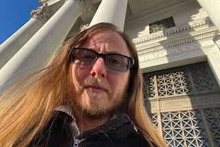 The author appears to be looking down at the audience. He has long brown hair, sunglasses, a beard, and a mustache tinged with white showing his age. Wrinkles are furrowing his forehead showing that he’s serious. Behind him are the pillars of a courthouse. An iron door is behind him. Above it are Greek letters. The sky is a striking blue in the background.