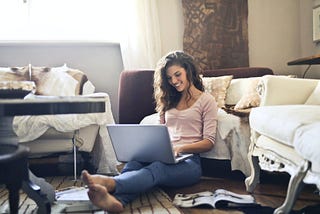 A smiling woman sits on the floor. She is surrounded by couches and has her laptop in her lap.