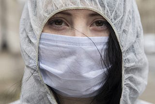 A young woman wears a face mask over her mouth & nose, as well as a protective hood over her head. She looks into the camera.