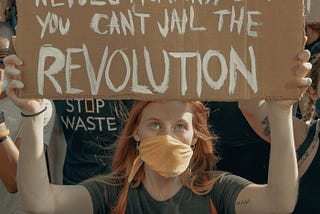 Photo of a young woman in a crowd of protesters marching together. She is wearing a COVID mask and holding up a cardboard sign with the hand painted message, “YOU CAN JAIL THE REVOLUTIONARY, BUT YOU CAN’T JAIL THE REVOLUTION”