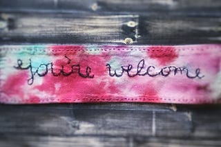 Photograph of a tie dyed piece of fabric with the words “You’re welcome” stitched on it. It was ironed onto a cloth mask.