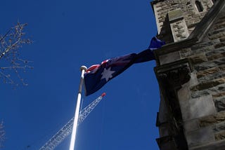 Australian Flag flying against blue sky with an old sandstone building and building crane in background.