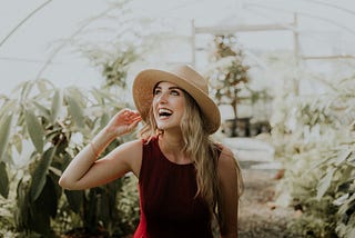 Blonde woman with sun hat on smiling and looking up while standing in a greenhouse. Rebecca Murauskas. Work-Life Integration Coach. Work-Life Balance. Freedom from overwhelm.