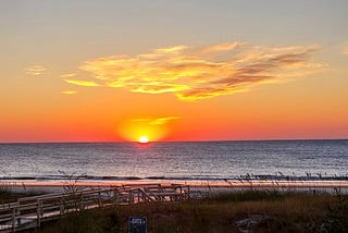 Author’s photo of a beach scene with a walking bridge in the setting sun filled with brilliant red, orange, and yellow.