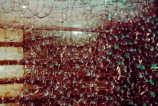 Close-up of cracked safety glass, fractured into hundreds of small, jagged segments, with warm and cool tones reflecting through the surface. Light filters through faintly, distorted and fragmented.