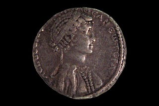 Egyptian or Roman coin depicting a silhouette of Cleopatra, with an elaborate braided hairstyle, pronounced nose and chin, and embellished clothing.