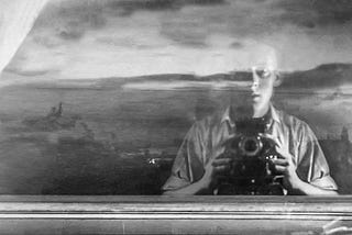 A 1947 black and white self-portrait of a young man, holding a camera, taking his own picture, his face reflected in a window with a cloudy sky beyond