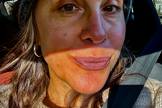 Photo of woman in hat with gray hair. Half her face is in a shadow, mouth in the light, half smiling