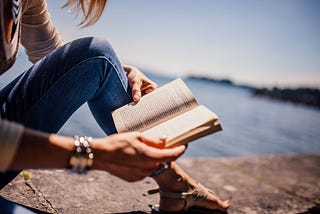 Photo of a woman reading a book looking out over a lake