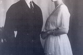 Felix and Irina standing together. He’s in a dark suit with short, slicked-back hair. She’s in a white day dress with pearls.