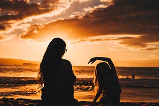 A woman and a young girl sitting on the beach, facing the sunset with their backs to us