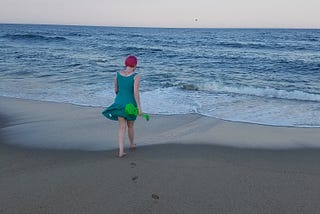 Photo of the author from behind, holding a ukulele and walking towards the waves on a beach.