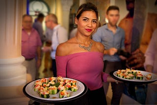 Image by ZigarrenZone on Pixabay of a Cuban woman serving food to male tourists in Cuba