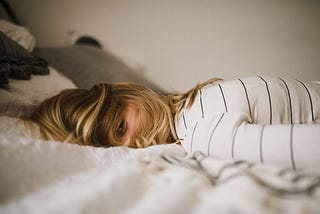 A woman laying forward on a bed, facing the camera with hair over her face.