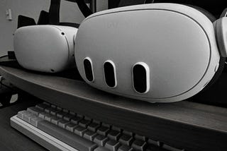 A Meta Quest 3 VR headset and its three camera bumps appear dominant in the frame on a desk, with a Quest 2 headset set to the left side. A keyboard sits below them on a desk tray.