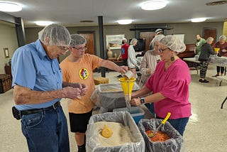 Several people at a workstation at a Rise Against Hunger packing event. A young man is pouring soy protein through a funnel into a plastic bag, being held by a woman. All three are wearing hairnets.