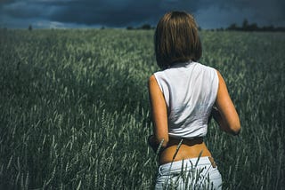 back view of girl with brown hair wearing white top and pants standing in grassy field looking at stormy sky