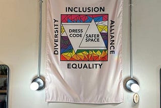 Dress Code Project flag — White Flag hung down from the ceiling with a square box with Inclusion on top, Alliance to the right, Equality at the bottom and Diversity on the left. Inside the box is rainbow colors plus pinks and whites and two triangles one upside down with the words Dress Code Safer Space.
