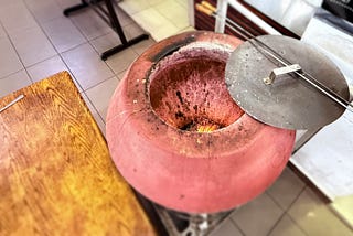 A traditional clay tandoor oven with an open lid, showing a fire inside, used for baking or grilling in a restaurant kitchen.