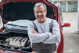 Smiling man with arms folded leaning on red car with open bonnet.