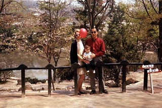 A photo from 1968 of me as an infant with my parents in a park. We’re in front of flowering trees and a pond, leaning against a wooden fence. My father is wearing a red shirt and black pants, my mother is wearing a white dress with a black coat, and a red balloon floats in front of her face. They are wearing sunglasses.