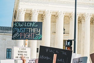 Image of protestors in front of the Supreme Court with signs reading “How long must they fight. Roe Wade,” “Fuck the abortion ban,” and “I love someone who had an abortion.”