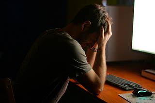 A distressed man in a dark room in front of a computer with their head in their hands.