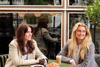 Two women are sitting at a bistro table drinking coffee.