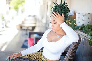 Black person with green locs seated outside on chair, possibly wondering why white people are still trying to do this hair this way and get away with it.