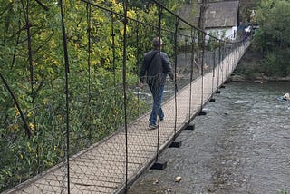 A man wearing a leather jacket and denim jeans walks across the boards of a suspension bridge over a river. The bridge is narrow with chain-fence sides, and trees are at the edge.