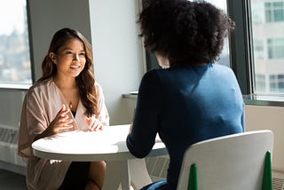 two people talking, sitting at a table in front of a window